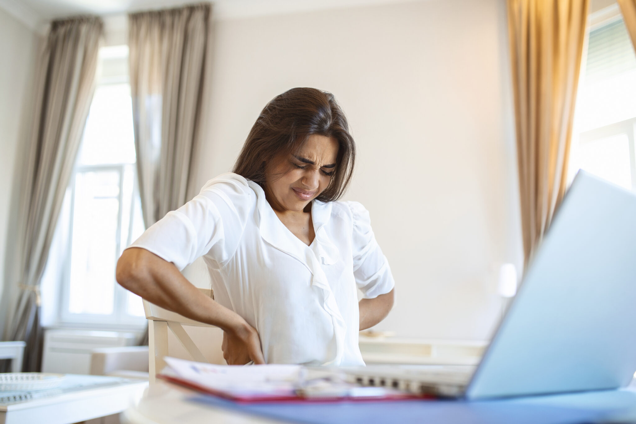 portrait of young stressed woman sitting at home office desk in front of laptop touching aching back with pained expression suffering from backache after working on laptop  scaled
