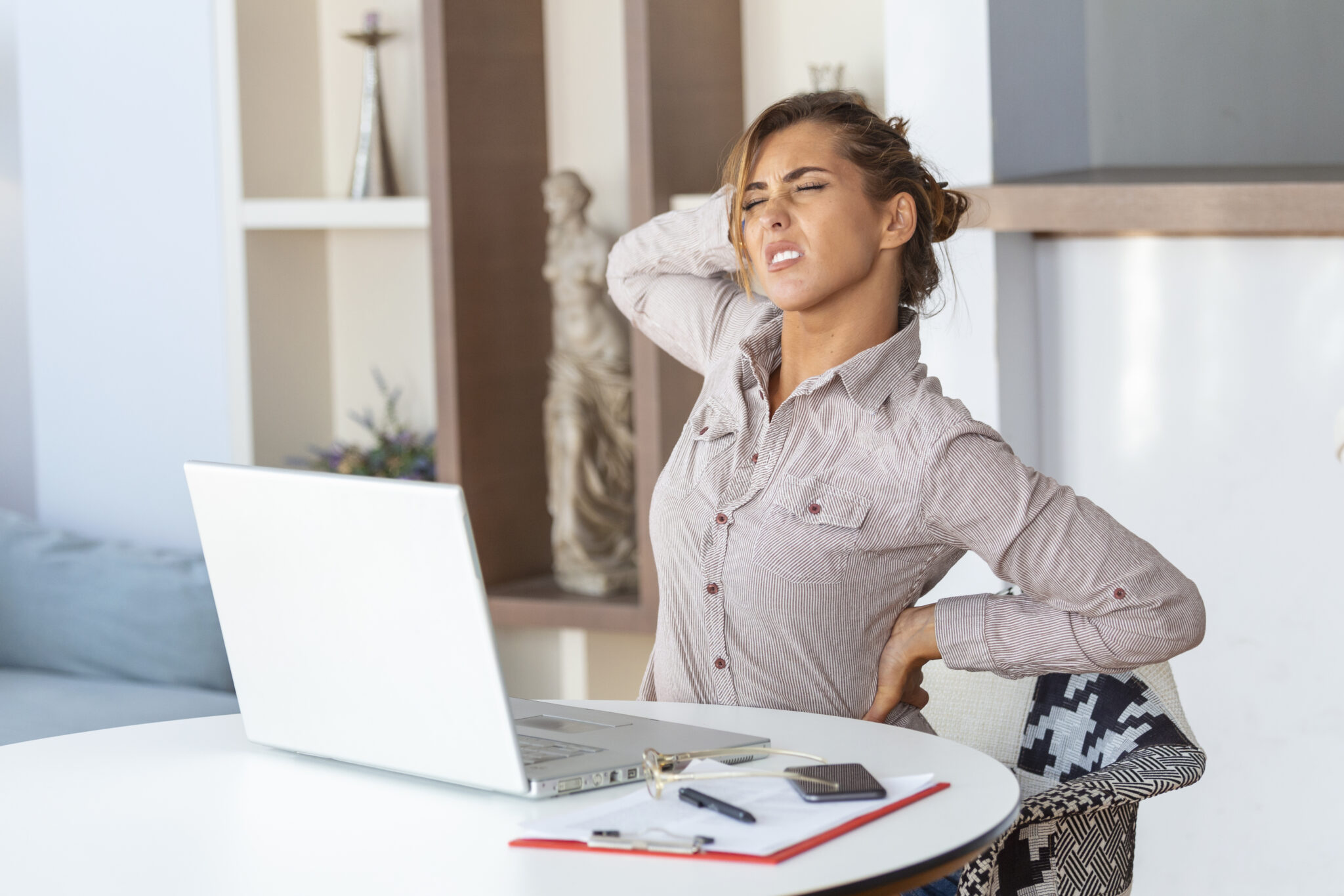 portrait of young stressed woman sitting at home office desk in front of laptop touching aching back with pained expression suffering from backache after working on laptop scaled